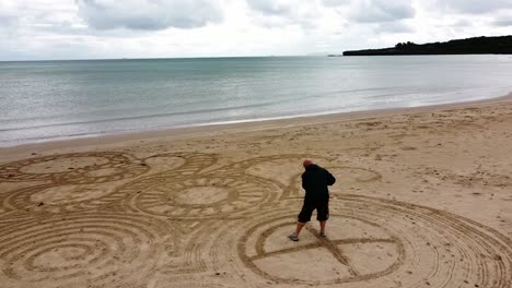 Luftaufnahme-Eines-Umkreisenden-Mannes,-Der-Am-Windigen-Walisischen-Goldenen-Strand-Ein-Interessantes-Zen-Sand-Kunstdesign-Kreiert