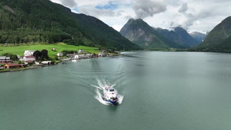 Tourist-boat-departing-Fjaerlandsfjorden-with-village-and-Boyabreen-glacier-seen-in-background-mountain-landscape
