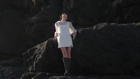 Eine-Atemberaubende-Frau-In-Einem-Weißen-Kleid-Mit-Eckigem-Ausschnitt-Und-Transparenten-Ärmeln-Schlendert-Anmutig-Und-Elegant-über-Die-Felsen-Am-Strand