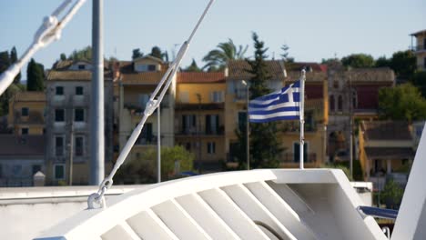Die-Griechische-Flagge-Wurde-Am-Bug-Des-Schiffes-Gehisst,-Mit-Hafenstadtgebäuden-Im-Hintergrund-Auf-Der-Insel-Korfu