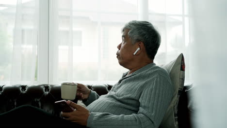 Un-Anciano-Activo-Que-Se-Relaja-En-Casa-Trabajando,-Usando-Un-Auricular-Bluetooth-Disfruta-Escuchando-Música-Desde-Un-Teléfono-Móvil-Sentado-Cómodamente-En-Un-Sofá