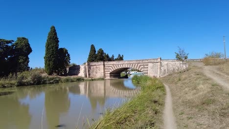 Puente-Sobre-El-Canal-Du-Midi-En-Le-Somail-Francia-Senderos-A-Ambos-Lados-Del-Canal-Conducen-Al-Pueblo-Turístico-En-Una-Mañana-De-Verano-Muy-Cálida