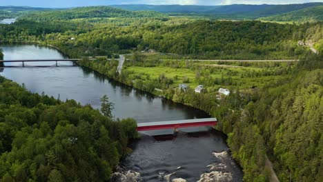 Backward-aerial-dolly-panning-up-to-reveal-covered-bridge-nestled-in-a-river-valley-in-a-hilly-green-landscape-in-Wakefield-Quebec-Canada