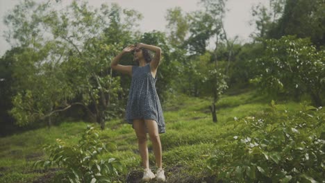 Full-body-woman-lifts-hands-to-fix-hair-while-standing-in-orchard-with-grassy-hill
