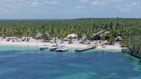 Cinematic-aerial-view-showing-coral-reef,-clear-water,-yachts,-sandy-beach-with-tourist-and-green-Tropical-landscape-during-sunny-day---Catalina-Island,-Dominican-Republic