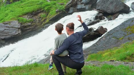 Son-sitting-on-fathers-lap-in-front-of-beautiful-Norway-river-at-Vikafjellet-mountain---Gestures-with-arm-to-show-how-big-the-river-is---Handheld-slow-motion