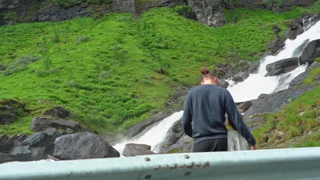 Father-and-son-watching-Sendefossen-river-along-road-to-Vikafjellet-Norway---Slow-motion-with-road-fence-in-foreground