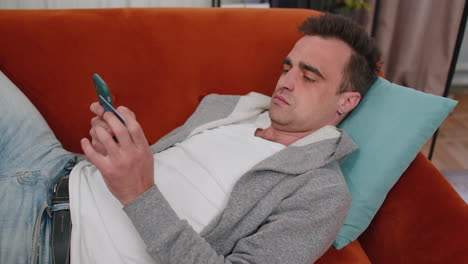 Man-lying-on-couch-using-credit-bank-card-and-smartphone-while-transferring-money-online-shopping