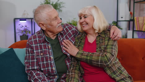 Happy-old-senior-elderly-family-couple-hugging,-laughing,-smiling-looking-at-camera-at-home-sofa