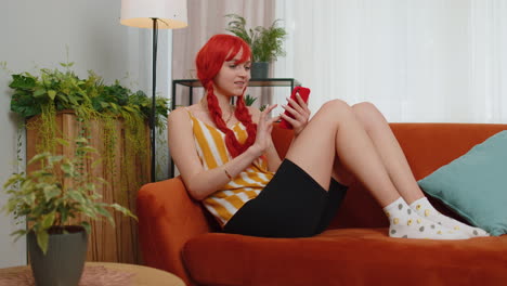 Young-ginger-girl-at-home-couch-use-smartphone-typing-browsing-say-wow-yes-found-out-great-big-win
