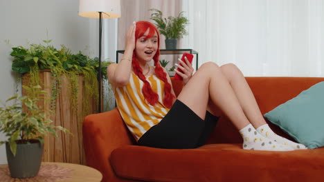 Young-ginger-girl-at-home-couch-use-smartphone-typing-browsing-say-wow-yes-found-out-great-big-win
