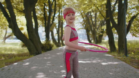 Athletic-fitness-toddler-girl-training-playing-twisting-Hula-hoop-circle-ring-around-waist-in-park