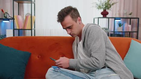 Man-sitting-on-couch-using-credit-bank-card-and-smartphone-while-transferring-money-online-shopping