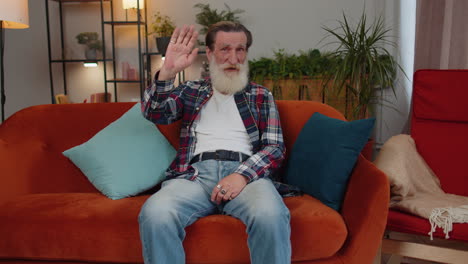 Senior-man-smiling-friendly-at-camera,-waving-hands-gesturing-hello,-hi,-greeting-at-home-on-couch