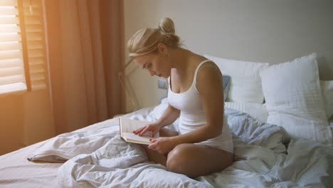 Lovely-young-woman-in-white-tank-top-and-panties-sitting-on-comfortable-bed-and-reading