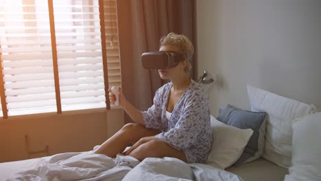 Young-woman-in-shirt-sitting-on-comfortable-bed-and-using-VR-headset-with-controller