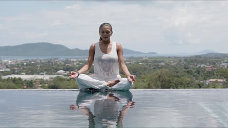 Mujer-Tranquila-Practicando-Yoga-En-Postura-De-Loto-Junto-A-La-Piscina-Contra-La-Ciudad-Turística