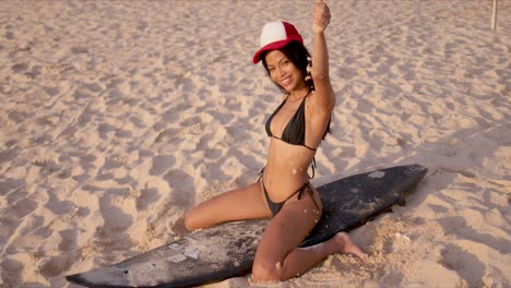 Cheerful-woman-in-bikini-sitting-on-surfboard-on-sand