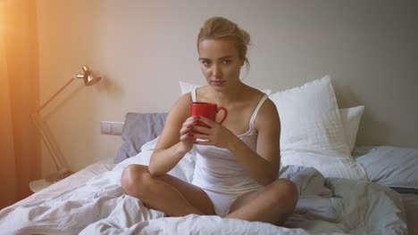 Pretty-woman-in-white-tank-top-and-panties-sitting-on-bed-and-enjoying-hot-beverage