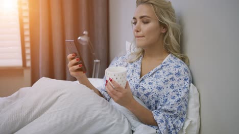 Beautiful-young-woman-browsing-smartphone-and-holding-mug-of-hot-beverage-sitting-on-bed