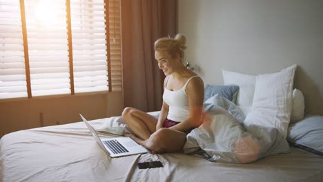 Charming-woman-in-pajamas-sitting-on-bed-near-laptop-and-video-chatting-with-her-friends