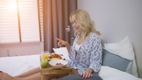 Attractive-young-woman-in-shirt-having-breakfast-while-sitting-on-bed-with-tray