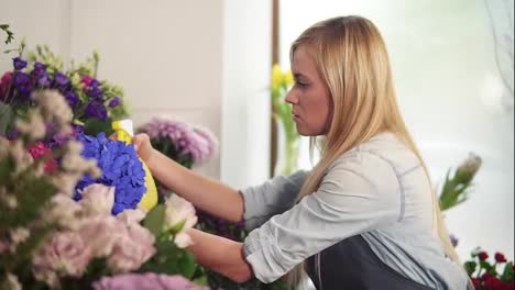 Young-attractive-blonde-female-florist-spraying-water-on-bunch-of-flowers-in-flower-shot.-Slow-Motion-shot