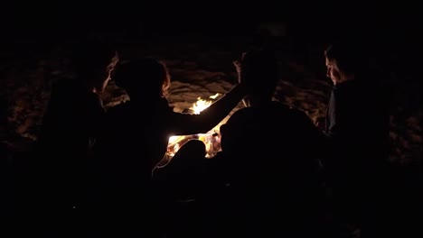 Back-view-of-young-people-sitting-together-by-the-fire-late-at-night-and-embracing-each-other.-Cheerful-friends-talking-and