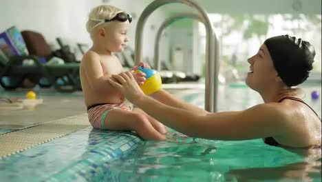 Young-mother-is-teaching-her-cute-blonde-toddler-to-swim-in-the-swimming-pool.-She-is-sitting-by-the-water-then-she-helps-him-to