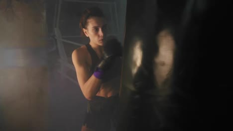 Caucasian-strong-female-kickboxer-exercising-with-a-boxing-bag-in-dark-gym-with-smoke-and-kicking-a-boxing-bag-with-her-hands
