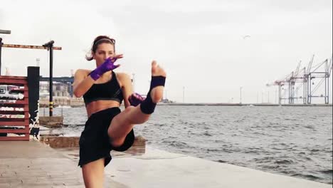 Young-female-boxer-practising-sidekick-of-her-legs-by-the-sea-in-slow-motion.-Beautiful-female-boxer-training-on-the-beach-in