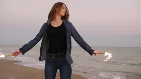 Young-beautiful-woman-running-by-the-sea-during-sunset-and-holding-burning-sparkling-candles-in-both-hands,-turning-around