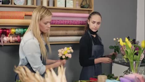Attractive-blonde-woman-in-apron-standing-with-her-coworker-at-counter-in-floral-shot-while-arranging-flowers
