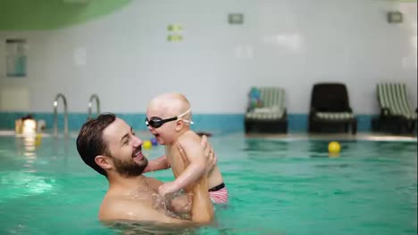 Young-father-lifting-his-little-boy-in-protective-glasses-from-the-water-while-teaching-him-how-to-swim-in-the-swimming-pool