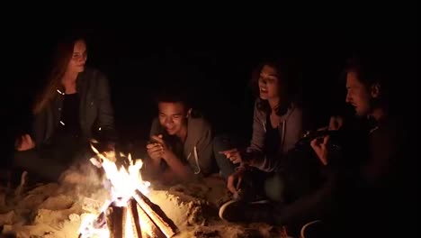 Round-camera-movement:-Multiracial-group-of-young-boys-and-girls-sitting-by-the-bonfire-late-at-night-and-singing-songs-and