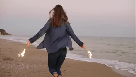 Back-view-of-young-attractive-woman-running-by-the-sea-during-sunset-and-holding-burning-sparkling-candles-in-both-hands