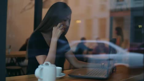 Blick-Von-Außen-Auf-Eine-Attraktive-Junge-Frau,-Die-In-Einem-Café-Telefoniert-Und-Dabei-Auf-Den-Bildschirm-Ihres-Laptops-Schaut