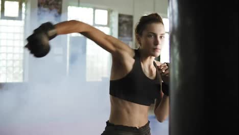 Young-caucasian-female-boxer-hitting-the-boxing-bag-with-her-hands-in-gloves-in-the-gym-with-smoke.-Tough-power-training-of-a