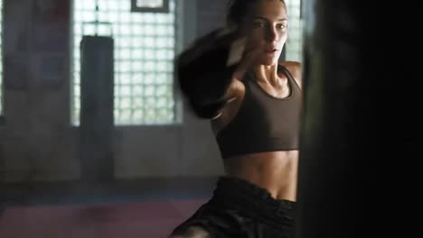 Strong-athletic-female-kickboxer-hits-a-boxing-bag-with-her-leg.-She-is-exercising-with-a-boxing-bag-in-dark-gym-with-smoke