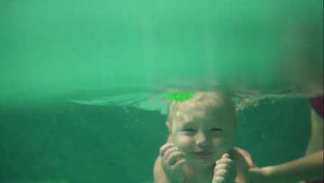 Cute-blonde-toddler-is-diving-under-the-water-in-the-swimming-pool-to-get-his-toy-while-his-mother-is-teaching-him-how-to-swim