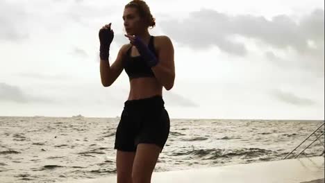 Athletic-karate-woman-makes-sidekick-of-her-legs-against-the-son-by-the-sea-in-slow-motion.-Beautiful-female-boxer-training-on