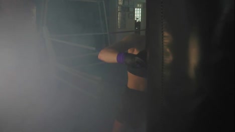 Slow-Motion-shot:-Strong-athletic-female-kickboxer-exercising-with-a-boxing-bag-in-dark-gym-with-smoke-and-kicking-a-boxing-bag