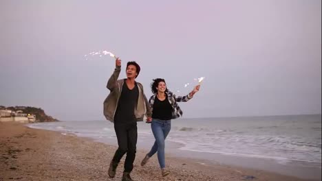 Young-happy-multi-ethnic-couple-holding-burning-sparkling-candles-and-running-by-the-sea-during-sunset.-Slow-Motion-shot