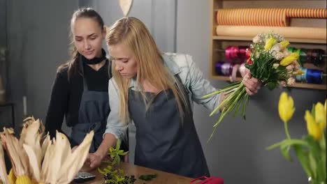 Blonde-florist-in-apron-with-her-coworker-at-counter-in-floral-shot-while-arranging-counting-the-price-for-bunch-of-flowers