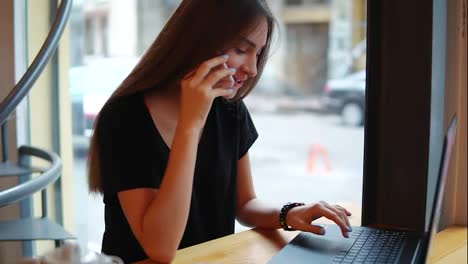 Attractive-young-woman-talking-on-the-phone-in-a-cafe-working-with-a-laptop-during-her-coffee-break.-Smiling-woman-sitting-by