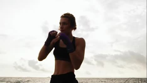 Athletic-young-woman-shadowboxing-by-the-sea-against-the-son.-Beautiful-female-boxer-training-on-the-beach-in-the-morning