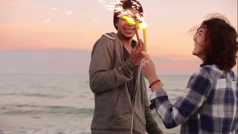 Cheerful-smiling-multi-ethnic-couple-holding-burning-sparkling-candles-standing-by-the-sea-during-sunset