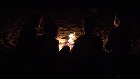 Back-view-of-unrecognizable-people-sitting-together-by-the-fire-late-at-night.-Cheerful-friends-talking-and-having-fun-together