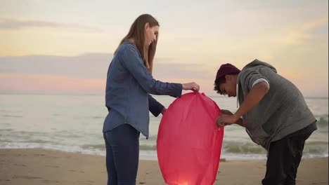 Young-multi-ethnic-couple-lightning-red-paper-lantern-before-launching.-Romantic-date-on-the-beach.-Attractive-woman-holding