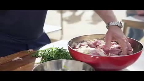 Man-is-mixing-meat-pieces-with-spices-and-onion-in-metal-bowl.-The-wooden-surface-outside.-Barbeque-time.-shot-in-4k